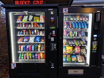 Zwei Snack-O-Matik Außenautomaten nebeneinander, gut gefüllt mit Snacks und Getränken